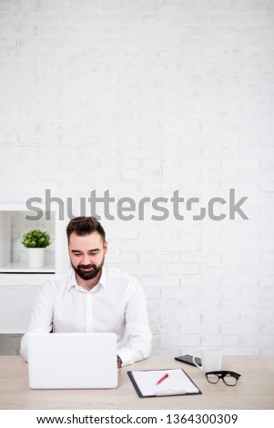 portrait of cheerful bearded businessman using laptop in office - copy space over white brick wall background