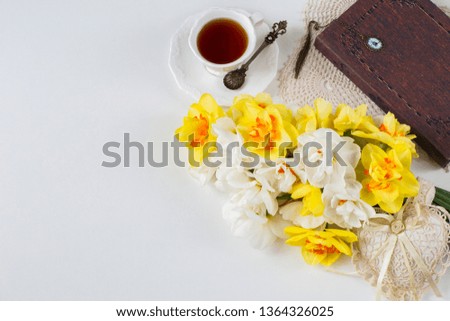 on a white table, a cup of tea, a book, a bouquet of daffodils and a lacy heart. Free space for text