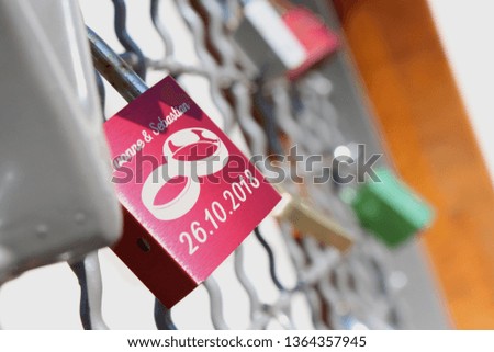 pink love lock hanging at a grid, foreign love locks in the background, big rings symbol in the middle of the lock, two names at top, date at the bottom