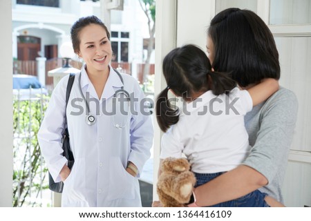 Asian female doctor in uniform standing near the entrance in the house and talking to mother about her little daughter Royalty-Free Stock Photo #1364416109