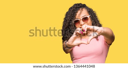 Young beautiful woman with curly hair wearing pink sunglasses smiling in love showing heart symbol and shape with hands. Romantic concept.