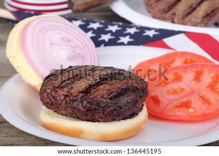 Closeup of a grilled hamburger on a bun with sliced onion and tomato and an American flag in background