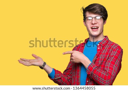 Young handsome student man wearing glasses over isolated background amazed and smiling to the camera while presenting with hand and pointing with finger.