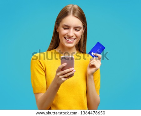 Happy girl looks at the phone and holds credit card. Photo of beautiful girl in yellow sweater on blue background. Emotions and pleasant feelings concept