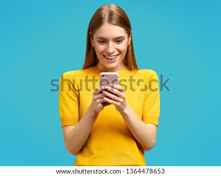 Happy girl with surprised expression looks at the phone. Photo of girl in yellow sweater on blue background.