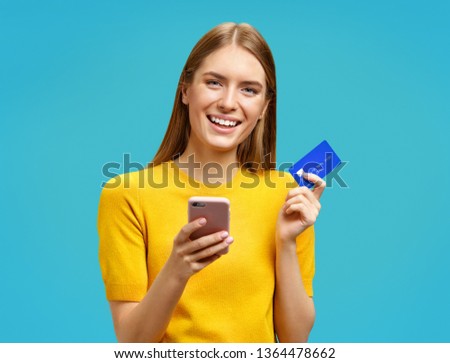 Smiling girl holds smartphone and credit card. Photo of beautiful girl in yellow sweater on blue background. Emotions and pleasant feelings concept.