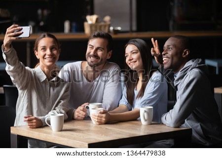 Biracial girl holding in hand phone make selfie photo with friends spend free time with diverse intimates in cafe, generation and modern wireless technology leisure activities in public place concept
