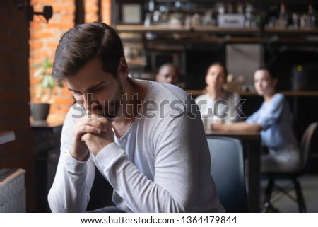 On foreground sad caucasian millennial guy outcast sitting separately from other diverse friends in cafe feels unhappy having communications difficulties, diffidence, jealousy, discrimination concept Royalty-Free Stock Photo #1364479844