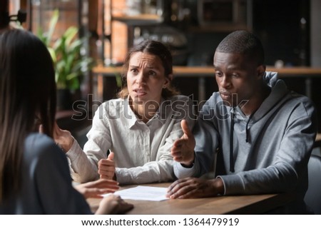 Dissatisfied couple customers having claim about contract conditions, diverse clients sitting at table disputing with agent about mortgage loan or real estate problem, fraud and bad contractor concept Royalty-Free Stock Photo #1364479919