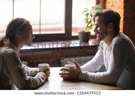 Side view smiling biracial woman sitting at table in cafe with caucasian man couple talking in cozy coffeeshop drinking tea coffee. Heterosexual friends romantic relationships or speed dating concept Royalty-Free Stock Photo #1364479952