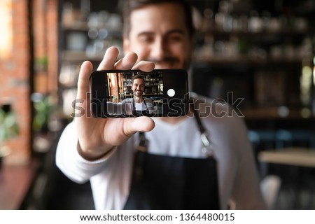Happy man wearing apron holds smartphone with camera on, waiter or cafe owner make selfie photo focus on device with self portrait image on mobile screen, share life in social media having fun concept