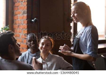 Diverse friends gathered together sitting in cafe talking with worker service waiting staff place make order food drink, young waitress business owner listening client wishes writing list in notepad Royalty-Free Stock Photo #1364480045