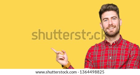 Young handsome man with a big smile on face, pointing with hand and finger to the side looking at the camera.