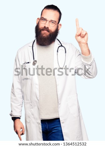 Young blond doctor man with beard wearing medical coat showing and pointing up with finger number one while smiling confident and happy.