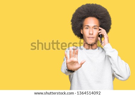 Young african american man with afro hair talking on smartphone with open hand doing stop sign with serious and confident expression, defense gesture