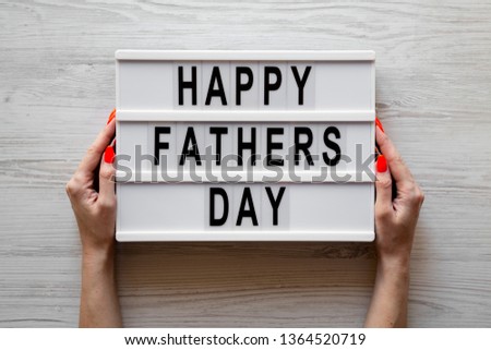 Female hands hold lightbox with 'Happy Fathers Day' words over white wooden background, top view. Overhead, from above, flat lay. 