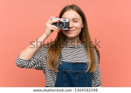 Young woman with overalls over pink wall holding a camera