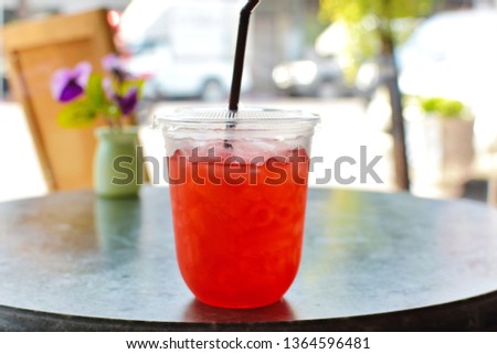 Ice tea with rose syrup and lemonade in grab and go glass with lid and plastic straw on wooden table in coffee shop blurred traffic view outside, summer beverage or plastic container is pollution 