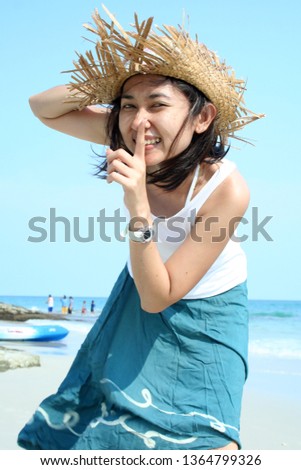 Simple Asian girl on beach smiling wearing straw hat making hand signal don't tell secret 