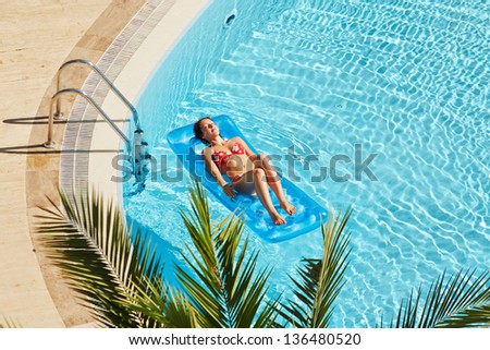 Young woman in red swimsuit sunbathes lying on inflatable mattress on water near to edge of pool