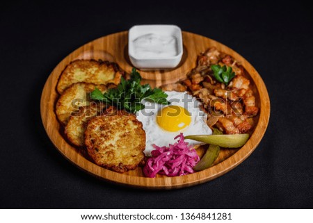 Potato pancakes with a fried egg, bacon, sourcream, horse radish, pickles and parsley on a wooden plate on black background