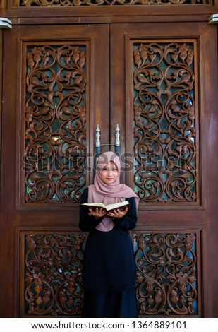Young malay muslim woman stand against carved wooden door while reading the holy quran.