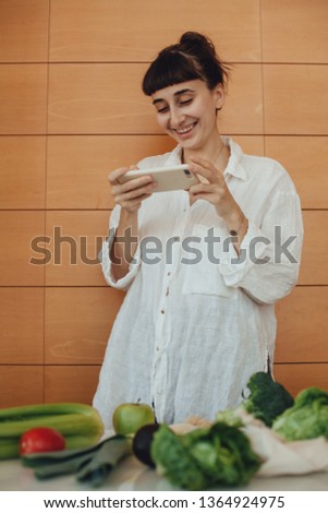Happy vegan girl making picture on her mobile phone of fresh vegetables and fruit that she bought in a cotton tote bag in eco grocery store, concept of no plastic and waste shopping and lifestyle