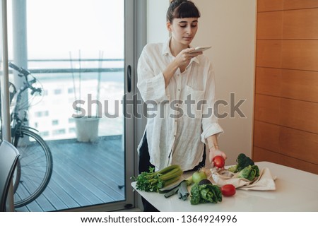 Pretty hipster young girl taking photo on her telephone camera of fresh green farm vegetables and fruit that she ordered online via delivery app, sustainable shopping and no plastic lifestyle