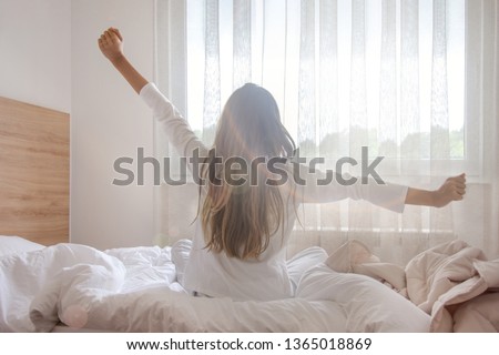 Young woman waking up in her bedroom, sitting on the bed stretching arms by the window Royalty-Free Stock Photo #1365018869