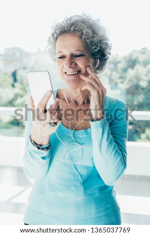 Joyful senior lady in earphones chatting with grandchildren. Grey haired woman standing at window indoors, holding smartphone and smiling at screen. Video call concept