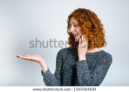beautiful curly woman advertises something on the palm, red-haired, isolated on gray background