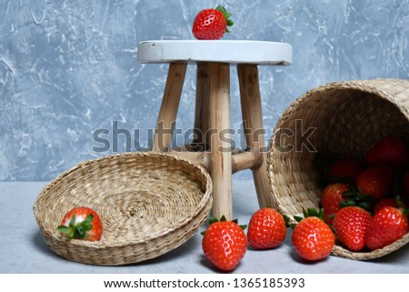 An image of a group of strawberries, a wooden round stand on three legs, a wicker basket and a lid to it on a white and gray surface on a white and gray-blue background