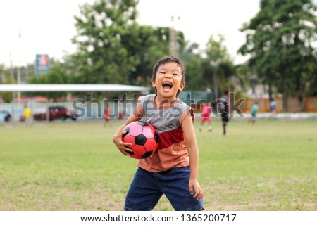 Happy Asian child boy laughing and smiling. He playing and holding a red ball in his hands at the football field. Kid and sports concept. Exercise for health. 