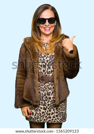 Beautiful middle age elegant woman wearing sunglasses and mink coat doing happy thumbs up gesture with hand. Approving expression looking at the camera with showing success.