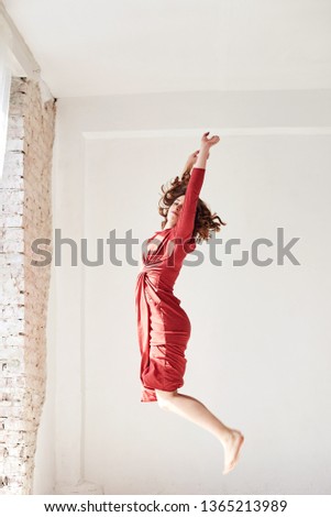 A full-length portrait of young pretty brunette woman with short curly hair, wearing red dress, jumping in the air, isolated on white background in light room 
