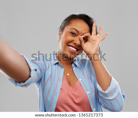 people and gesture concept - happy african american young woman taking selfie looking through fingers over grey background