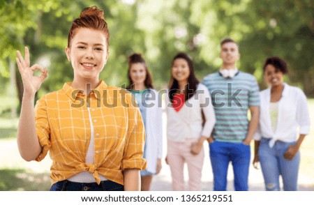 gesture and people concept - smiling red haired teenage girl in checkered shirt showing ok hand sign over group of friends at summer park background