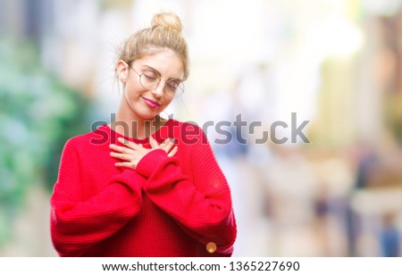 Young beautiful blonde woman wearing red sweater and glasses over isolated background smiling with hands on chest with closed eyes and grateful gesture on face. Health concept.