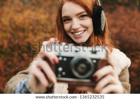 Lovely redheaded young girl listening to music, holding photo camera while standing outdoors