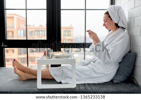 A girl in a bathrobe photographs her breakfast on a tray near the window on a soft window-sill.
