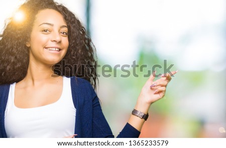 Young beautiful girl with curly hair with a big smile on face, pointing with hand and finger to the side looking at the camera.