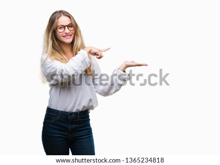 Young beautiful blonde business woman wearing glasses over isolated background amazed and smiling to the camera while presenting with hand and pointing with finger.