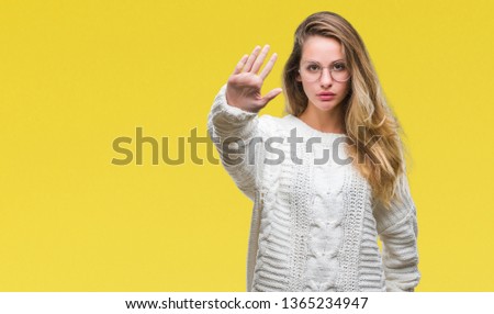 Young beautiful blonde woman wearing winter sweater and sunglasses over isolated background doing stop sing with palm of the hand. Warning expression with negative and serious gesture on the face.