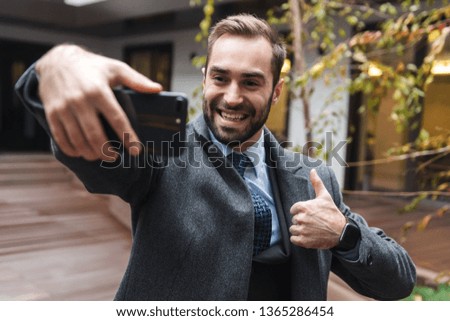 Attractive smiling young businessman wearing suit walking outdoors, taking a selfie