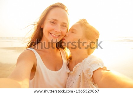 Happy stylishly mother kiss daughter and taking selfie at sandy beach on a sunset.mothers Day.little girl blonde and beautiful woman taking pictures on the phone tourism abroad online video calling