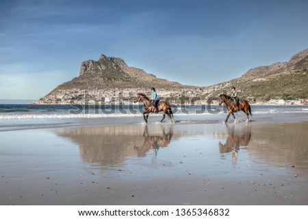 Two teenage girls on horseback cantering in the water at low tide on the beach under a cloud sky with a beautiful mountain in the background and reflections of themselves in the water on the shore