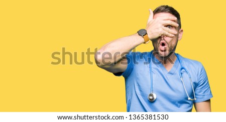 Handsome doctor man wearing medical uniform over isolated background peeking in shock covering face and eyes with hand, looking through fingers with embarrassed expression.