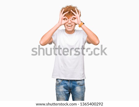 Young handsome man with afro hair wearing casual white t-shirt doing ok gesture like binoculars sticking tongue out, eyes looking through fingers. Crazy expression.