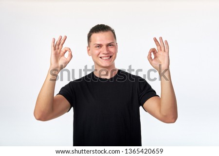 Attractive cherrful young man dressed in black shirt showing OK gesture, smiling and looking at camera. Positive feelings. Hand gesture all good. I'm okay. Studio shot on white wall background.