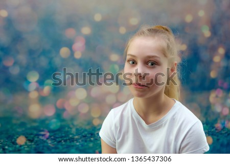 A concept portrait of a cute pretty blonde teen girl with long hair is sitting in a white T-shirt on a colorful background in the studio and talking. She's right in front of the camera.